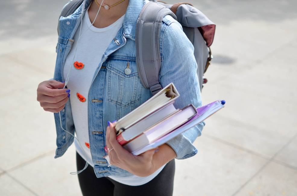 Person wearing denim jacket with ear buds in and wearing a backpack and holding books