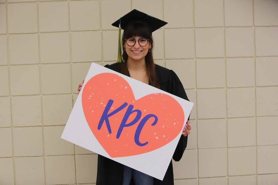 Woman wearing classes and graduation cap and gown holding a KPC sign