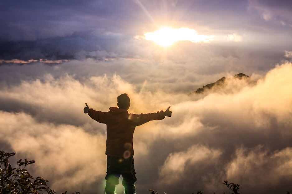 Man on mountain giving thumbs up looking at clouds