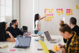 Group of people around a conference table while someone points to sticky notes on a wall