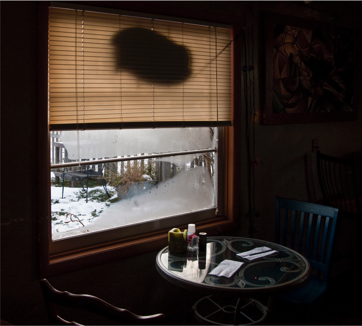 A table with two chairs, set against a frosty window, the shade half draw the dark outline of a turned off closed sign, the table is set with salt and paper shakers, a small glass dish with suger and artifical sugar, a piller candle and two napkins the view outside is of a dusting of snow covering a green ground, planter, small picket fence and metal chair 